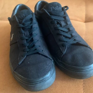 Svarta sneakers från Polo Ralph Lauren - Svarta sneakers från Polo Ralph Lauren med klassisk låg profil. Skorna har snörning och är tillverkade i textil med brun sula. Polo-logga syns på sidan och på sulan. Perfekta för en stilren och enkel look. Använda ett fåtal gånger 5-6