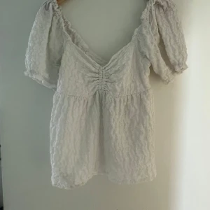 Vit blus med puffärmar och knytning i ryggen - Säljer en söt vit blus med strukturmönster, puffiga korta ärmar och v-ringning. Blusen har rynkdetalj framtill och knytning i ryggen för extra detalj. Perfekt till sommaren!