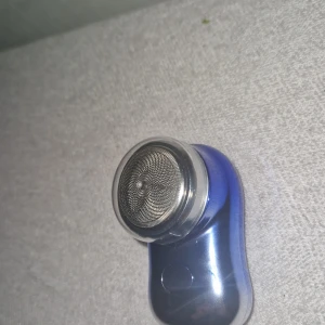 Elektrisk ansiktsrengörare i blått - Elektrisk ansiktsrengörare med rund metallisk borste och blått handtag. Kommer med ett genomskinligt plastlock. Perfekt för att applicera toner och rengöra huden på djupet.