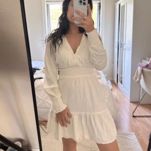 Vit långärmad klänning med volang från Zara - Superfin vit klänning från Zara med v-ringning, smock i midjan och långa ärmar med resår vid ärmslut. Klänningen har volangdetaljer nertill och är tillverkad i ett lätt material som passar perfekt till vår och sommar.