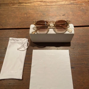 Solglasögon - Snygga solglasögon från CHIMI med transparent beige båge och bruna tonade glas. Rektangulär form och diskret logga på skalmen. Perfekt accessoar för soliga dagar. Nypris 1300