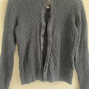 Grå kabelstickad kofta - Mysig blå kofta med kabelstickat mönster och knappar framtill. Långärmad modell med ribbade muddar vid ärmslut och nederkant. Perfekt att slänga på sig över en t-shirt eller blus.