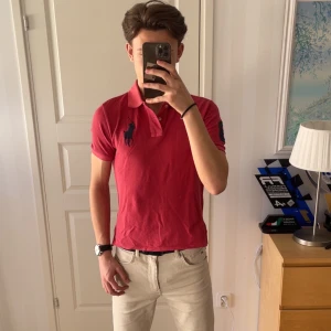 Röd pikétröja från Ralph Lauren - Snygg röd pikétröja från Ralph Lauren med klassisk krage och knappar. Tröjan har en broderad logga på bröstet i mörkblått. Perfekt för en stilren look.