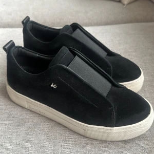 Svarta sneakers från KCobler - Stilrena svarta sneakers från KCobler med vit platt sula och elastiskt band över vristen istället för snörning. Ovandelen är i mocka och skon har en minimalistisk design med diskret logga på sidan. 