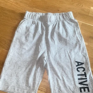 Grå träningsshorts från Luca & Lola - Säljer ett par ljusgrå träningsshorts från Luca & Lola med trycket 'ACTIVE' längs ena benet. Shortsen har resår i midjan och är tillverkade i mjuk bomull, perfekta för sport eller chill. Längden är ca50 cm