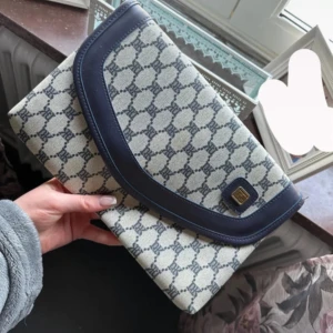 Mönstrad kuvertväska med blå detaljer - Säljer en snygg kuvertväska med blått och vitt mönster samt mörkblå kant och detaljer. Väskan har ett klassiskt monogrammönster och stängs med lock. Invändigt finns en liten ficka med dragkedja. Perfekt accessoar för att lyfta din outfit.