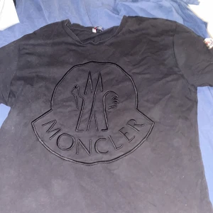 Svart t-shirt från Moncler med stor logga - Säljer en svart t-shirt från Moncler i storlek M. T-shirten har en stor, broderad Moncler-logga på bröstet och en mindre logotyp på ärmen. Klassisk rund hals och korta ärmar. Perfekt för dig som gillar stilrena märkesplagg.