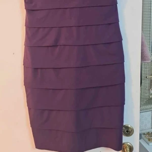 Lila klänning med rosettdetaljer från En Focus Studio - Säljer en lila klänning med v-ringning och vackra rosettdetaljer upptill. Klänningen har en figurnära passform och lager på lager-design nedtill. Perfekt för dig som vill sticka ut med en elegant look.