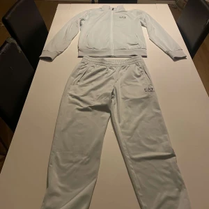 Vit träningsdress från Emporio Armani EA7 - Säljer en stilren vit träningsdress från Emporio Armani EA7. Setet består av en zip-jacka och matchande byxor med elastisk midja. Båda plaggen har EA7-logga och är tillverkade i ett mjukt och lätt material.