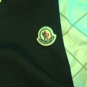 Svart Moncler pufferjacka med huva - Säljer en svart pufferjacka från Moncler med huva och dragkedja. Jackan har blanka partier på framsidan och ärmar i sweatshirtmaterial. Vit och svart randig detalj över bröstet samt Moncler-logga på ärmen.