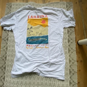 Vit t-shirt med Lahaina-print från Rip Curl. Där det står lahania är baksidan - Vit t-shirt från Rip Curl med färgglatt tryck på ryggen som föreställer Lahaina, Maui. Framsidan har ett litet tryck med palm och sol. T-shirten har normal passform och är tillverkad i mjuk bomull.