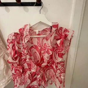 Blommig croppad blus med knyt - Säljer en blommig croppad blus i vitt och rött med volangdetaljer och knyt framtill. Perfekt för sommardagar när du vill sticka ut lite extra. Tunn och luftig modell med korta ärmar och v-ringning.