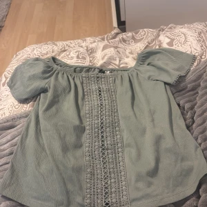 Grön blus med spetsdetalj - Säljer en ljusgrön blus med korta puffärmar och dekorativ spets längs hela framsidan. Blusen har en lös passform och är perfekt för vår och sommar. Superfin att matcha med jeans eller kjol!