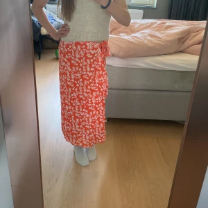 Röd blommig omlottkjol från ASOS - Säljer en röd omlottkjol från ASOS med vitt blommönster. Kjolen är lång, har volangdetaljer och knyts i midjan. Perfekt för dig som vill ha en färgstark och somrig look.