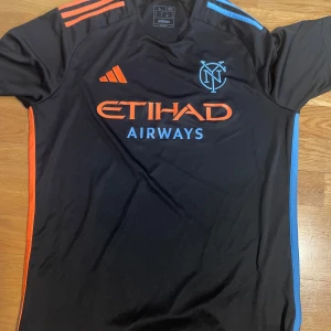 Svart NYCFC fotbollströja från Adidas - Säljer en svart fotbollströja från Adidas för New York City FC med orange och blå detaljer. Tröjan har korta ärmar, klubbmärke på bröstet och Etihad Airways som sponsortryck. Perfekt för fotbollsfans som gillar snygg design.