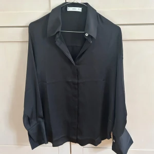 Svart blus/skjorta från Mango - Säljer en svart skjorta från Mango i storlek XS. Skjortan har klassisk krage, långa ärmar med breda manschetter och knappar framtill. Materialet är mjukt och har en lätt glansig finish💕Använt en gång