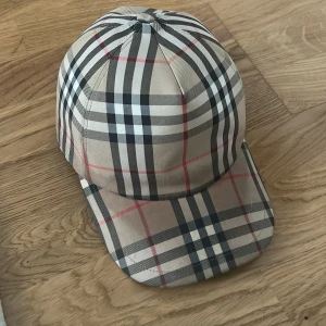 Beige rutig keps från Burberry - Säljer en klassisk beige keps från Burberry med det ikoniska rutiga mönstret i svart, vitt och rött. Kepsen har justerbar baksida och broderad Burberry-logga. Perfekt för dig som vill ha en stilren accessoar.