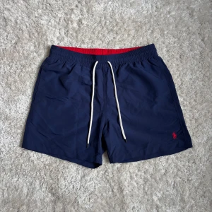 Polo Ralph Lauren badshorts - Snygga marinblå badshorts från Polo Ralph Lauren med vit snörning i midjan och röd insida | Perfekta för sommardagar vid stranden eller poolen | Size M, passar folk som har S som gillar lite längre benlängd | Inga defekter | Midjemåttet som mest utsträckt är 40cm 