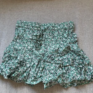 Grön blommig volangkjol från ZARA - Supersöt grön kortkjol med blommönster från ZARA. Kjolen har volanger och knytband i midjan för extra detalj. Har även shorts inuti i samma mönster💕