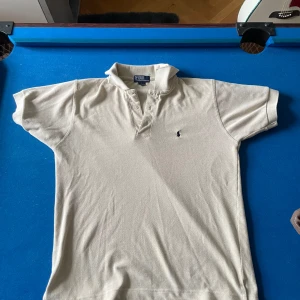 Beige  pikétröja från Polo Ralph Lauren - Klassisk beige pikétröja från Polo Ralph Lauren i 100% bomull. Tröjan har korta ärmar, krage och knappar framtill samt den ikoniska broderade loggan på bröstet. Perfekt för en stilren och tidlös look.