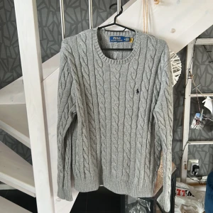 Grå kabelstickad tröja från Polo Ralph Lauren - Säljer en klassisk grå kabelstickad tröja från Polo Ralph Lauren. Tröjan har rund halsringning, lång ärm och den ikoniska broderade loggan på bröstet. Perfekt för lager-på-lager och enkel att matcha med det mesta. Vid några funderingar hör av dig // William 