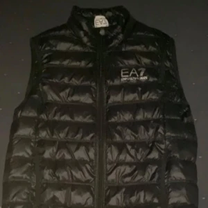 Svart dunväst från Emporio Armani EA7 - Snygg svart dunväst från Emporio Armani EA7 med quiltad design och dragkedja framtill. Västen har en hög krage och logga på bröstet. Perfekt för lager-på-lager och stilrena outfits.