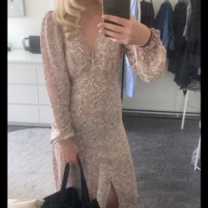 Blommig långklänning från BikBok - Supersöt långklänning från BikBok i beige med vitt blommönster. Klänningen har v-ringning, långa ärmar och en slits framtill. Perfekt för vår och sommar! Tillverkad i återvunnet material.