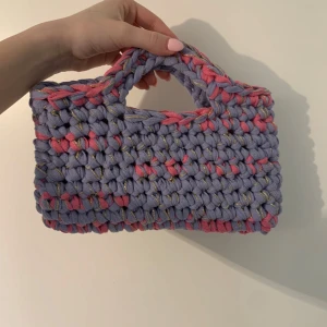 Handgjord lila och rosa virkad handväska - Superfin handväska i grovt virkat material med lila och rosa toner samt inslag av guldtråd. Väskan har ett rektangulärt format med inbyggda handtag och är perfekt för att bära småsaker. Unik accessoar som verkligen sticker ut! Väskan är 27cm bred och 15 cm hög