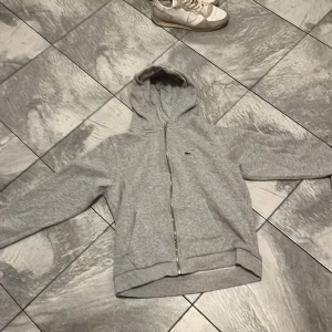 Grå hoodie med dragkedja från Lacoste - Säljer en stilren grå hoodie från Lacoste med dragkedja framtill och klassisk krokodillogga på bröstet. Tröjan har huva och ribbade muddar vid ärmslut och nederkant. Perfekt för en avslappnad look. Anledning varför jag säljer för liten!