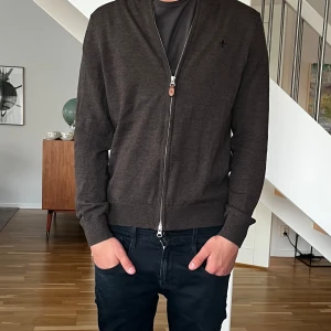 Mörkbrun kofta med dragkedja från Ralph Lauren - Säljer en stilren mörkbrun kofta från Ralph Lauren med hel dragkedja framtill. Tröjan har lång ärm, ribbade muddar och diskret logga på bröstet. Perfekt att slänga på sig över en t-shirt för en clean look.