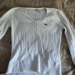 Vit kabelstickad tröja från Lacoste - Säljer en klassisk vit kabelstickad tröja från Lacoste med v-ringning och lång ärm. Tröjan har ribbade muddar och den ikoniska krokodilloggan på bröstet. Perfekt för en stilren och tidlös look.