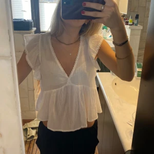 Vit blus med volang och spetsdetaljer - Säljer en söt vit blus med v-ringning och spetskant längs halsen. Blusen har korta volangärmar och är tillverkad i ett lätt, luftigt material. Perfekt för varma dagar och ger en romantisk känsla till outfiten.