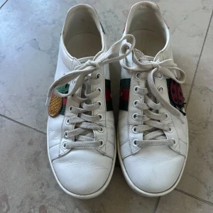Gucci sneakers med broderade detaljer - Säljer ett par vita Gucci sneakers i skinn med klassiska gröna och röda ränder på sidorna. Ena skon har en broderad ananas och den andra en broderad nyckelpiga. Baksidan har metallicröd och grön häl med Gucci-logga. Rund tå, snörning och platt sula. Perfekt för dig som gillar unika detaljer!