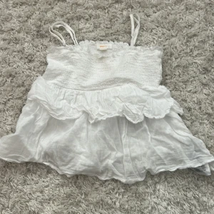 Vit volangtop från Gina Tricot - Supersöt vit linnetopp från Gina Tricot med smala axelband och smockad överdel. Toppen har dubbla volanger och luftig känsla, perfekt för varma dagar. Materialet är lätt och känns som bomull. Passar dig som gillar romantisk och somrig stil.