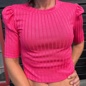 Ribbad rosa croppad topp från NA-KD - Säljer en ribbad croppad topp från NA-KD stl.S/XS  i stark rosa färg. Toppen har puffiga axlar, korta ärmar och rund halsringning. Den är figurnära och har en snygg struktur med vertikala ränder. 