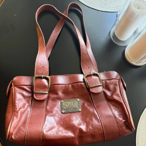 Brun axelväska från Dolce & Gabbana - Säljer en brun/vinröd vintage axelväska från Dolce & Gabbana i skinn med rejäla axelremmar och snygga metallspännen. Vet inte om den är äkta. Jag köpte den second hand.