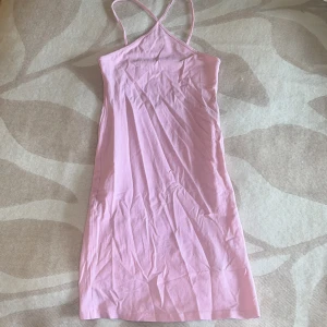 Rosa axelbandslös sommarklänning - Säljer en ljusrosa klänning med smala axelband som korsas i ryggen. Klänningen är kort och har en enkel, rak passform som ger en chill vibe. Perfekt för varma dagar och lätt att styla med sneakers eller sandaler.