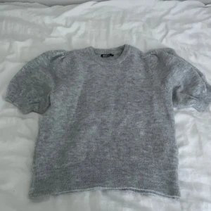 Grå stickad tröja med puffärm - Supermysig grå stickad tröja från Gina Tricot med korta puffärmar och rund hals. Tröjan har ribbade muddar vid ärmslut och nederkant, vilket ger en snygg och chill look. Perfekt att matcha med jeans eller kjol för en enkel men trendig stil.