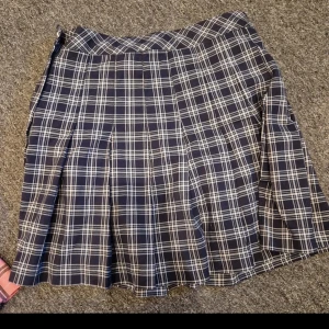 Marinblå rutig kortkjol - Säljer en snygg marinblå kortkjol med vitt rutmönster. Kjolen har klassiska veck och en skön passform som ger en cool och ungdomlig vibe. Perfekt för dig som gillar preppy stil och vill sticka ut med ett tidlöst mönster.