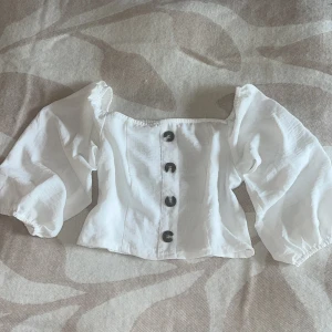 Vit croppad blus med puffärmar - Supersöt vit blus med croppad passform och stora puffärmar. Framsidan har fyra svarta knappar och offshoulder-design. Baksidan är smockad för extra komfort. Perfekt för dig som gillar trendiga och romantiska vibes.