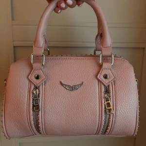 Zadig väska, rosa - Säljer en snygg rosa Zadig handväska i skinnimitation med silverfärgade nitar längs kanterna och coola dragkedjor fram och bak. Väskan har två stadiga handtag och man får med ett matchande axelband. Perfekt storlek för en utekväll eller till vardags.