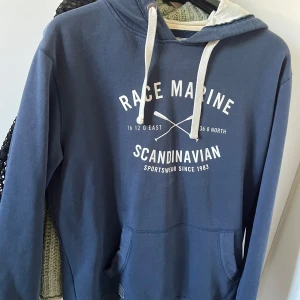 Blå hoodie  - Säljer en blå hoodie från Race Marine med vit text och tryck på bröstet. Tröjan har vit fodrad huva, snörning och en stor magficka. Perfekt för chill dagar och har en skön, mjuk känsla tack vare bomullsmaterialet. Klassisk streetstil med lång ärm och relaxed passform.