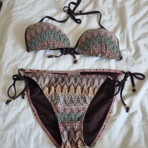 Mönstrad bikini från Athlecia - Snygg bikini från Athlecia med färgstarkt mönster i grönt, rosa, gult och brunt. Bikinin har knytband med små guldfärgade pärlor och en cool boho-vibe. Materialet är 92% polyester och 8% lurex som ger en lätt glittrig effekt. Perfekt för strandhäng och poolparty!