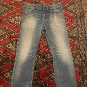 Blå Diesel jeans med slitningar - Säljer ett par snygga ljusblå jeans från Diesel med slitna detaljer och klassisk femficksdesign. Jeansen har en rak passform och tydliga tvättade partier framtill och baktill. Perfekta för dig som gillar en avslappnad och trendig look.