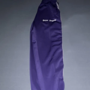 Palm Angels lila trackpants M - Säljer ett par lila trackpants från Palm Angels med vit logga på vänster sida och vita stripes längs benen. Byxorna har elastisk midja och är gjorda i 100% polyester för en skön och sportig vibe. Perfekt för dig som gillar streetwear och vill sticka ut.