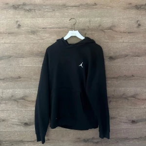 Svart hoodie från Jordan med logga - Svart hoodie från Jordan med den klassiska Jumpman-loggan i vitt på bröstet. Tröjan har huva, lång ärm och en stor magficka framtill. Mjuk insida och ribbade muddar vid ärmslut och nederkant. Perfekt för chill eller streetwear.