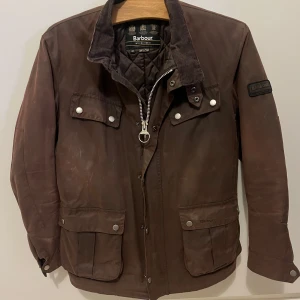 Barbour jacka  - Snygg brun jacka från Barbour International med quiltat foder och klassisk krage i manchester. Jackan har flera fickor med tryckknappar, dragkedja och Barbour-logga på ärmen. 
