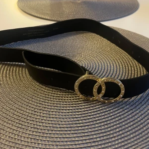 Svart skärp med gulddetaljer, 85 cm - Snyggt svart bälte i äkta skinn med två runda guldfärgade spännen dekorerade med små stenar. Bältet är stilrent och passar perfekt till jeans eller klänning för att lyfta din outfit. Tillverkat i Sverige.