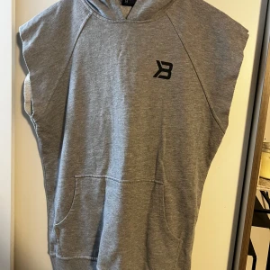 Grå ärmlös hoodie från Better Bodies - Säljer en grå ärmlös hoodie från Better Bodies i storlek S. Tröjan har huva, magficka och tryckt logga både fram och bak. Perfekt för gymmet eller chill dagar. Materialet är mjuk bomull med ribbad nederkant och råa kanter vid ärmarna för en sportig vibe.
