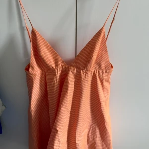 Randig orange klänning med smala axelband - Säljer en fräsch orange klänning med vita smala ränder och tunna axelband. Klänningen har v-ringad front och ärmlös design, perfekt för varma dagar. Lätt och luftig passform som ger en chill vibe. Storlek M.